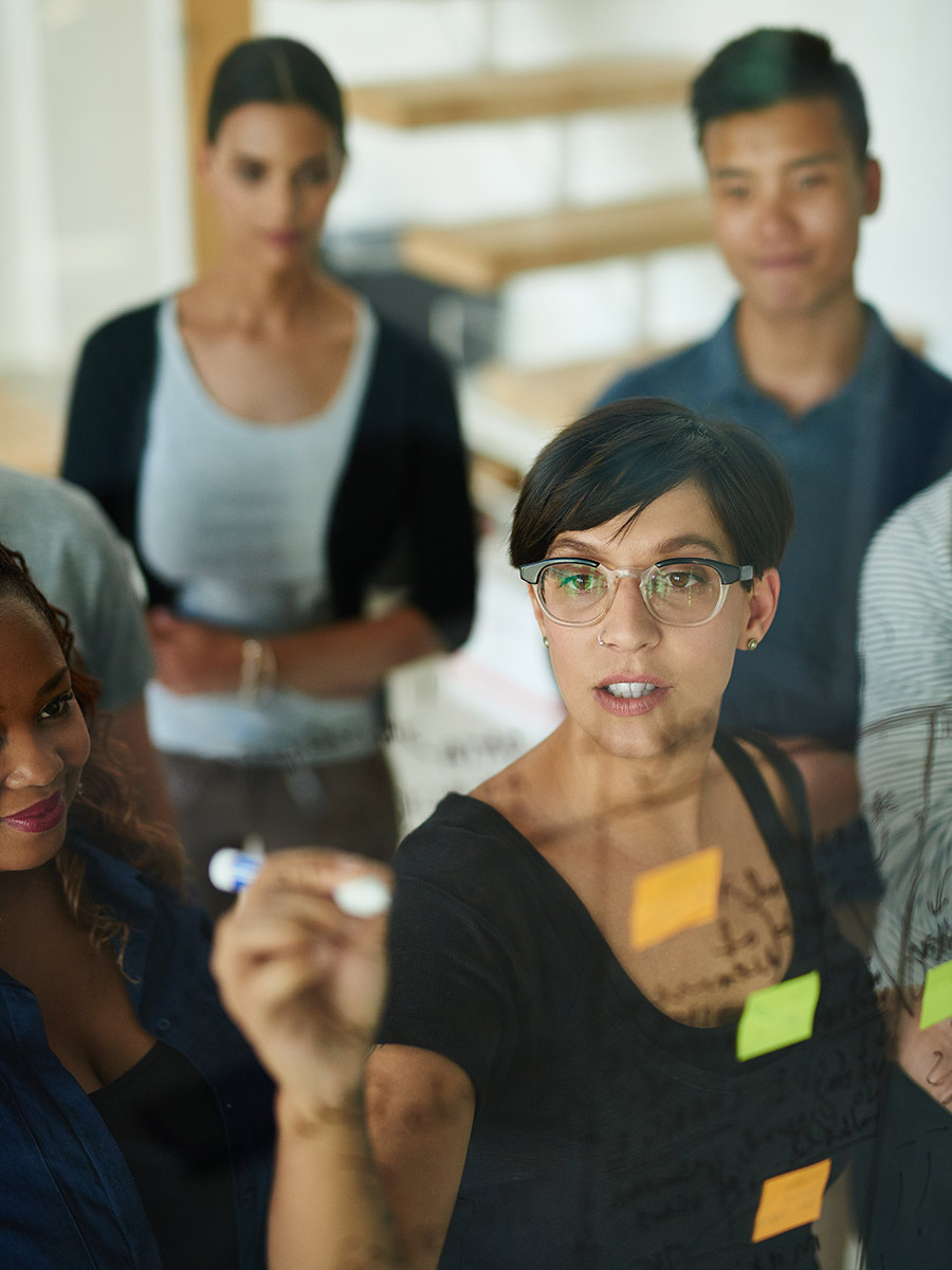 Woman writes on glass marker board with marketing team discussing buyer personas