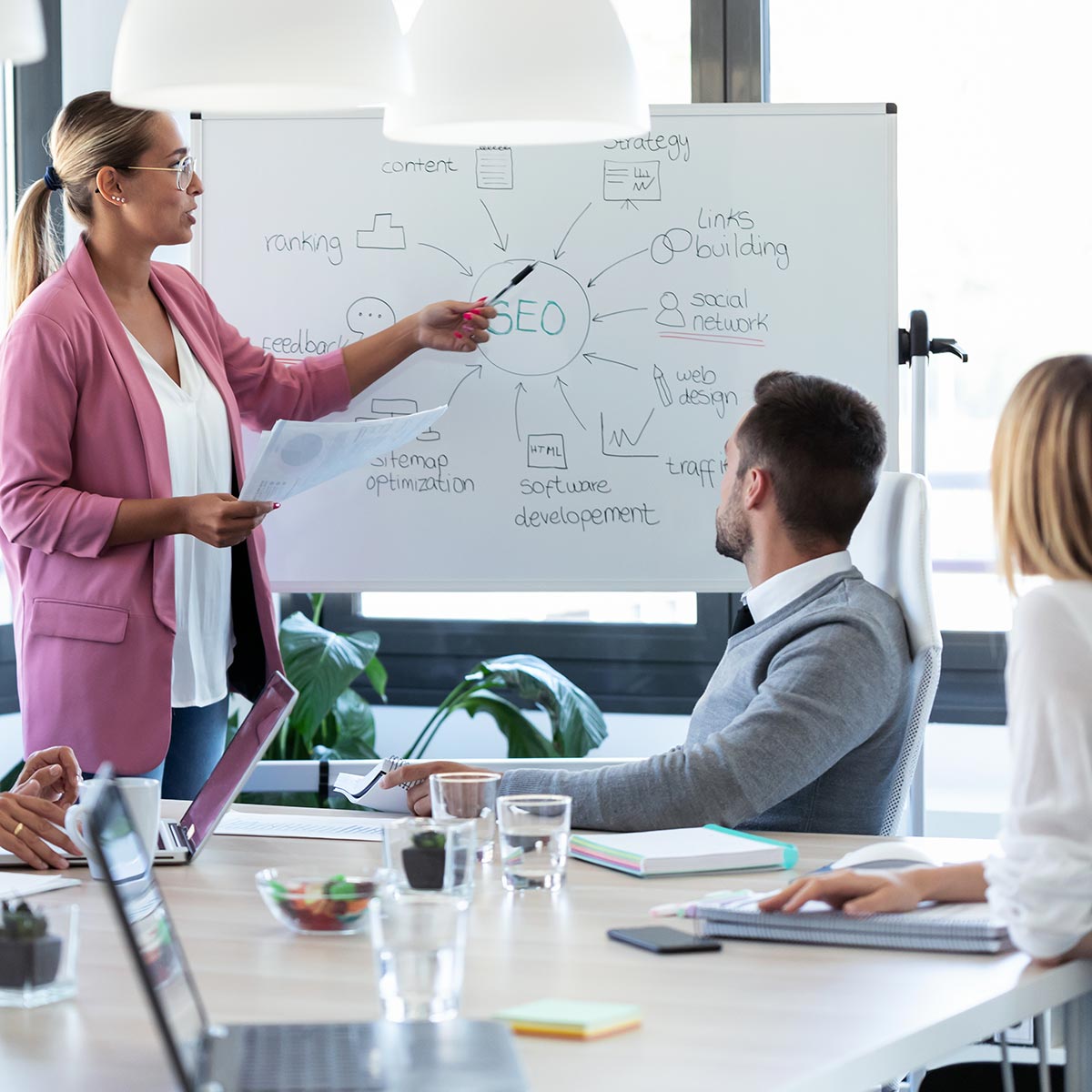 In a team discussion about SEO for manufacturers, a female team member points to a whiteboard with an SEO chart.